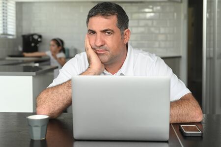 Upset father (age 40-50) looking at camera, using laptop as the pandemic coronavirus (COVID -19)...