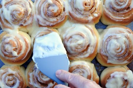 Person point of view adding cream on cinnamon roll cookies in kitchen.