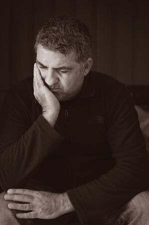 Upset mature adult man sitting in the dark looking away from camera isolated on black background.