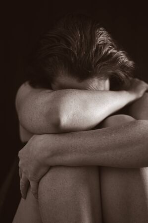 Sad adult woman sitting in the dark covering her face isolated on black background.の写真素材