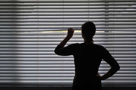 Suspicious adult woman looking outside home window from behind Venetian curtains. Concept photo of person that have Pistanthrophobia and Agoraphobia, a person who don't trust fear of leaving the house.の写真素材
