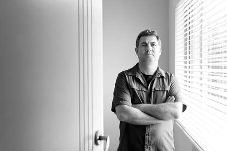 Upset adult man standing with arms folded in a bedroom looking at camera.
