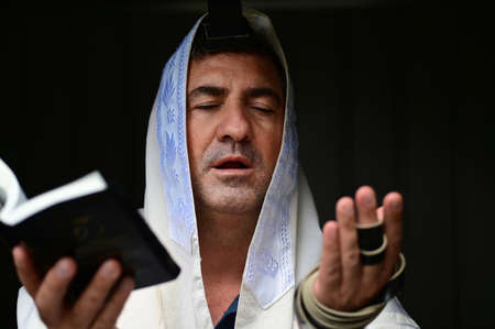 Adult Jewish man holding Siddur book (Jewish prayer book) , wearing tallit and tefillin (Jewish prayer clothing), paying the morning pray.Isolated on black background. Real people. Copy spaceの写真素材