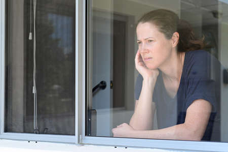 Upset middle aged adult  woman (age 30-40) looking through home window. Real people. Copy space