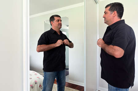 Middle aged man (age 40-50) looking at mirror buttoning up a shirt getting dressing up in home...