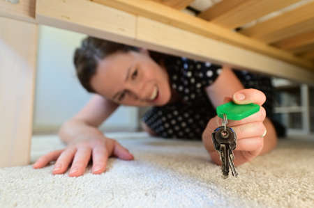 Happy adult woman (age 30-40) finding home keys under the bed in a domestic house bedroom. Real...