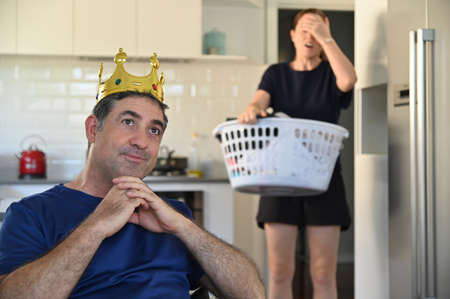 Lazy mature adult man (age 40-50) behaving like a king, watching TV while his wife upset carrying a laundry busket  in the background. Laziness concept. Real people. Copy spaceの写真素材