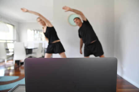 Shallow dept of field of adult couple (age 40-50) exercising at home living room watching an online body workout training video clip on a laptop screen. Real people. Copy spaceの写真素材