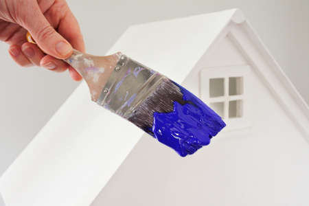Person holding a paint brush with blue paint color against a white house. Renovation, home decoration and painting house concept. No people. Copy spaceの写真素材