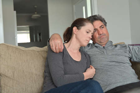 Upset couple (male age 40-50 and female age 30-40) sitting on a couch alone with puzzled face...