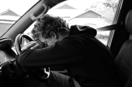 A sad and depressed mature adult man (male age 40-50) sitting in a car leaning on vehicle steering wheel outside his home.の写真素材