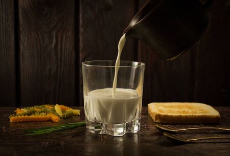 a glass of milk falling from a brown saucepan onto a wooden bottom with pasta and breadの写真素材