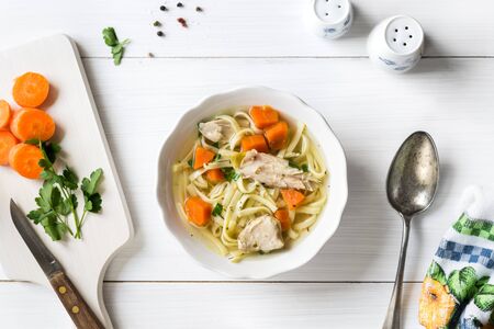 Fresh chicken soup with vegetables and pasta in a bowl with carrot and parsley on white table, top viewの写真素材