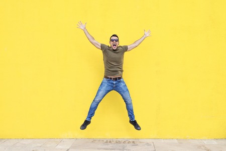 Front view of a young happy man wearing sunglasses jumping against a yellow bright wall in a sunny...