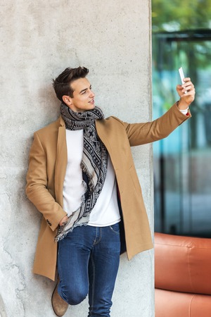 Side view of young trendy man leaning on a wall outdoors wearing denim clothes while taking a selfie in sunny dayの写真素材