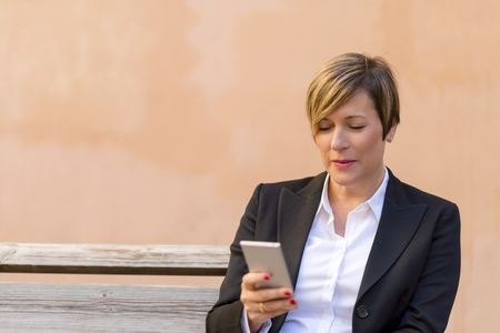 Front view of elegant smiling business woman sitting on a bench while using a mobile phone enjoying the conversation outdoorsの写真素材