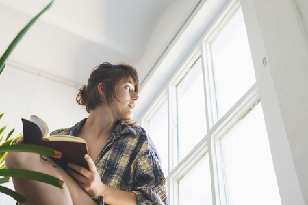 Low angle view of beautiful sensual woman wearing shirt with ponytail sitting on staircase at home while holding a book next to a window in a sunny dayの写真素材