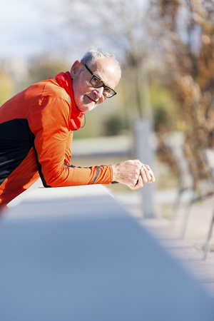 Front view of a senior runner man with sportswear sitting on a wooden fence while looking cameraの写真素材