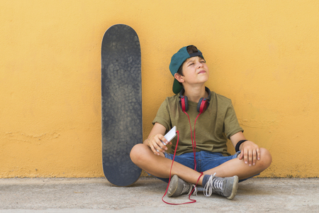 Front view of a young boy sitting on ground leaning on a yellow wall while using a mobile phone to listening musicの写真素材