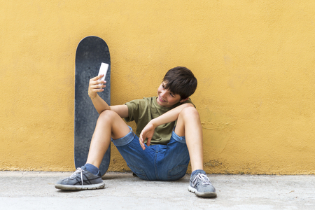Front view of a young boy sitting on ground leaning on a yellow wall while using a mobile phone to take a selfieの写真素材