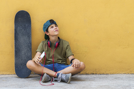 Front view of young boy sitting on ground leaning on a yellow wall while using a mobile phone to listening musicの写真素材