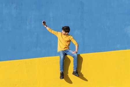 Front view of a young boy wearing casual clothes sitting on a yellow fence against a blue wall while using a mobile phoneの写真素材