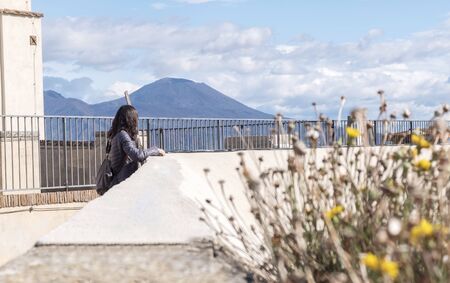 Rear view of thoughtful traveler woman looking away while standing by stoned fence against cloudy skyの写真素材