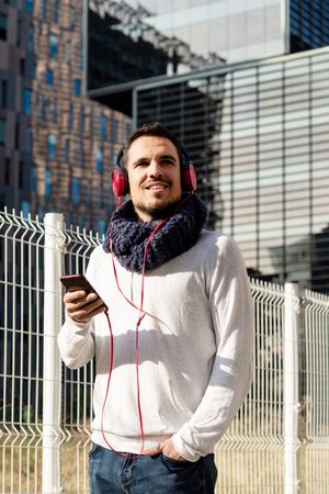 Young bearded man with headphones and holding smartphone while walking against skyscrapers in sunny...