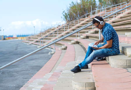 Young male with headphones and sunglasses sitting on stairs outdoors using tablet