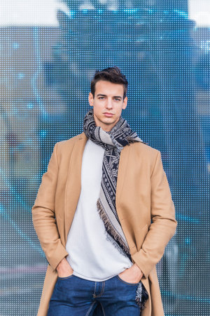 Portrait of stylish young man with coat standing outdoors and leaning on a led panelの写真素材