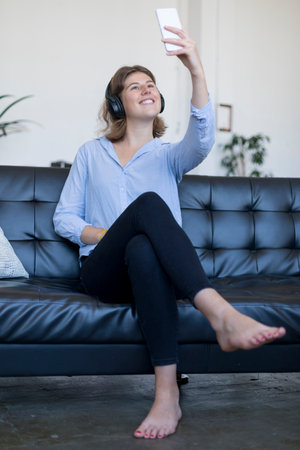 A Young smiling woman sitting on leather sofa with headphones taking a selfie