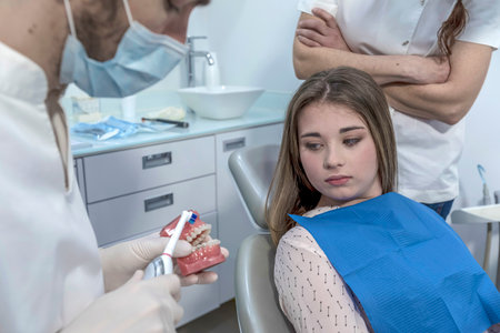 Young dentist in white uniform and protective medical mask showing jaw model with braces to female patient in dentistry office with modern equipment.の写真素材