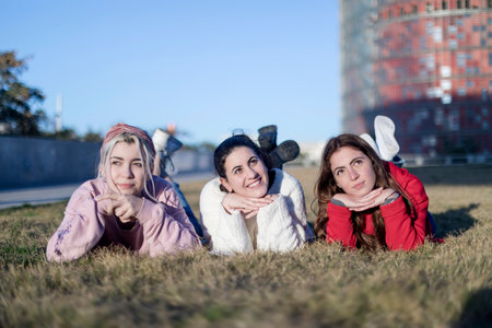 Three young women lying on their stomachs on the grass, posing playfully with hands under chins, with a clear sky and urban backgroundの写真素材