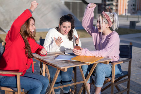 Three young women gather around a tablet and documents, engaged in a group study session outdoors. They're focused yet relaxed, with an urban backdrop.の写真素材