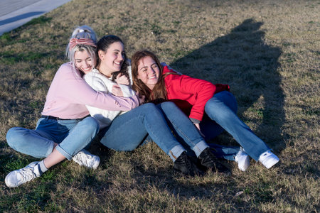 Three friends enjoy a relaxed moment sitting on the grass, basking in the sunlight, sharing smiles and a close bond in casual attireの写真素材