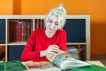 Young woman with bleached blonde hair, focusing on reading. Dressed in red, sitting at a library...