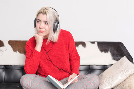 Young woman with blonde hair and red sweater, wearing headphones, sits cross-legged on a couch, holding a book and resting her chin on her hand, looking pensive.の写真素材