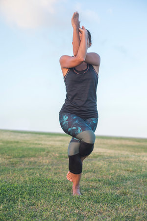 A woman in athletic wear performs the eagle yoga pose on a grassy field, balancing on one leg with arms crossed, against a clear blue sky background.の写真素材