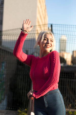 A cheerful young woman in a red sweater waves with a bright smile, enjoying the sunny urban setting, her platinum blonde hair glowing in the light.の写真素材