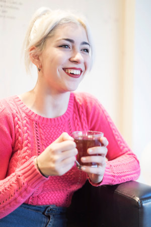A young woman in a pink sweater sips a hot drink, her expression content and relaxed in a warmly lit indoor environment, adding to the cozy ambianceの写真素材