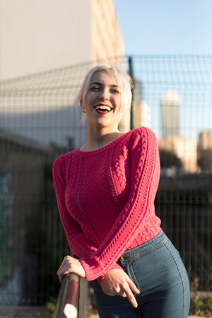 A joyful young woman in a vibrant red sweater leans on a railing outdoors, smiling and enjoying the sunlight, against a blurred urban backgroundの写真素材
