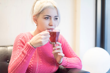 A young woman in a pink sweater sips a hot drink, her expression content and relaxed in a warmly lit indoor environment, adding to the cozy ambianceの写真素材