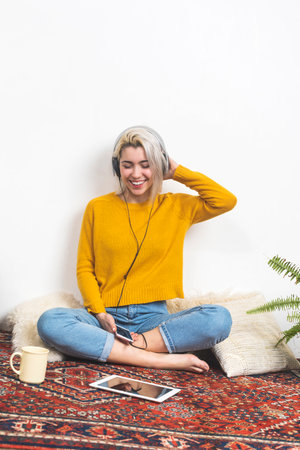 Caucasian blonde woman in a yellow sweater, smiling and sitting cross-legged, wearing headphones, holding a phone, enjoying music indoors.の写真素材
