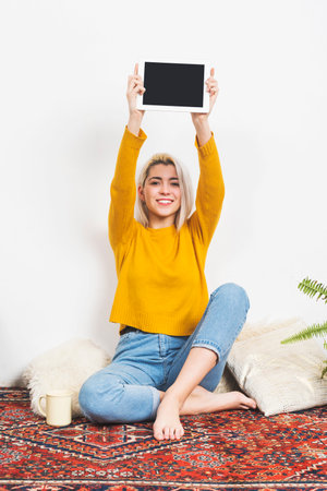 Caucasian woman with blonde hair in a yellow sweater holds a blank tablet aloft, sitting cross-legged on a decorative rug with cozy elements.の写真素材