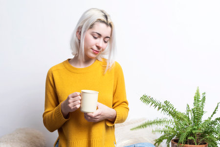 A Caucasian woman with blonde hair looks down softly, holding a beige mug. She wears a yellow sweater, sitting near a green plant, evoking calm vibes.の写真素材