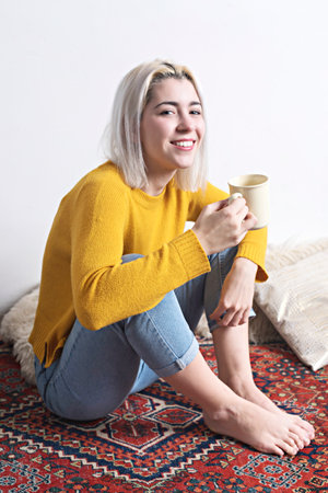 Caucasian woman with blonde hair, in a yellow sweater and jeans, smiling warmly while holding a mug. Relaxed and cozy atmosphere on a patterned rug.の写真素材
