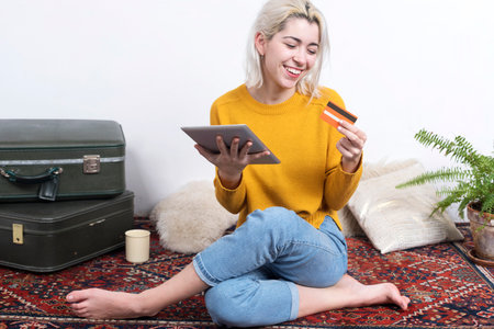 Caucasian woman sitting on a patterned rug, holding a tablet and credit card, smiling while shopping online. Vintage suitcases and plants in the background.の写真素材