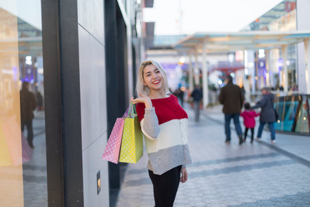 A happy Caucasian woman with blonde hair carries shopping bags on her shoulder in a stylish mall. She wears a trendy red sweater and looks cheerful.の写真素材