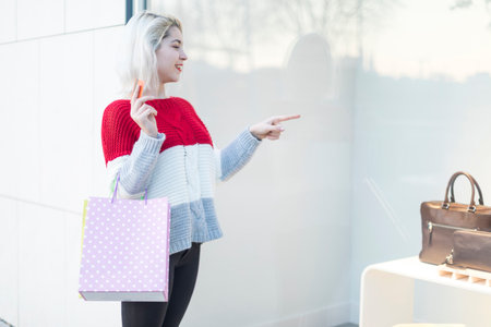 A young Caucasian woman wearing a red sweater points at a product display while holding a shopping bag and credit card, showcasing modern retail experiences.の写真素材