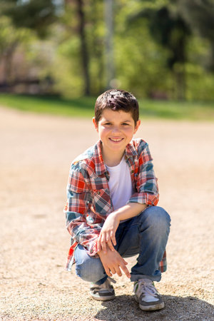 A young Caucasian boy with short dark hair, wearing a plaid shirt, jeans, and sneakers, squats on a gravel path in a sunlit park, looking curious.の写真素材